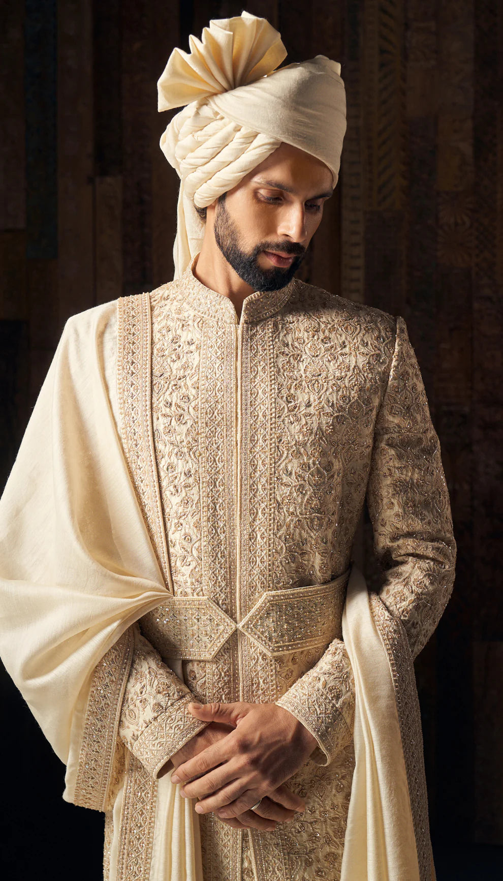 Man in almond-beige sherwani by designers Shyamal and Bhumika, against a dark backdrop