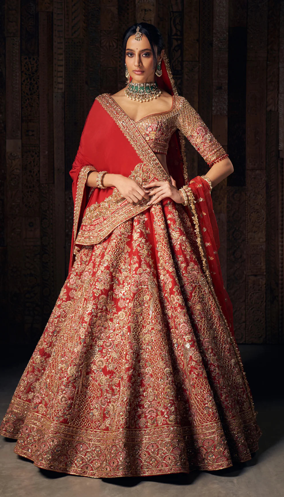 Woman in a bright red gold embroidered bridal lehenga by designers Shyamal and Bhumika, against a dark wooden backdrop