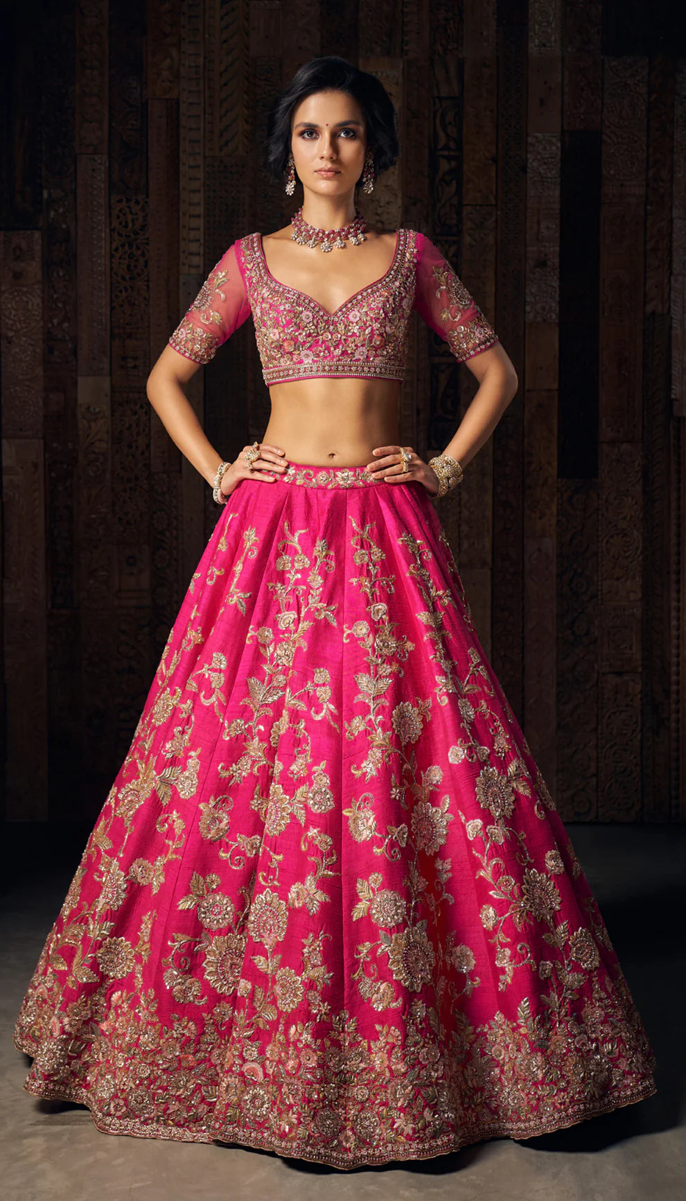 Woman in a bright pink gold floral  embroidered bridal lehenga by designers Shyamal and Bhumika, against a dark wooden backdrop