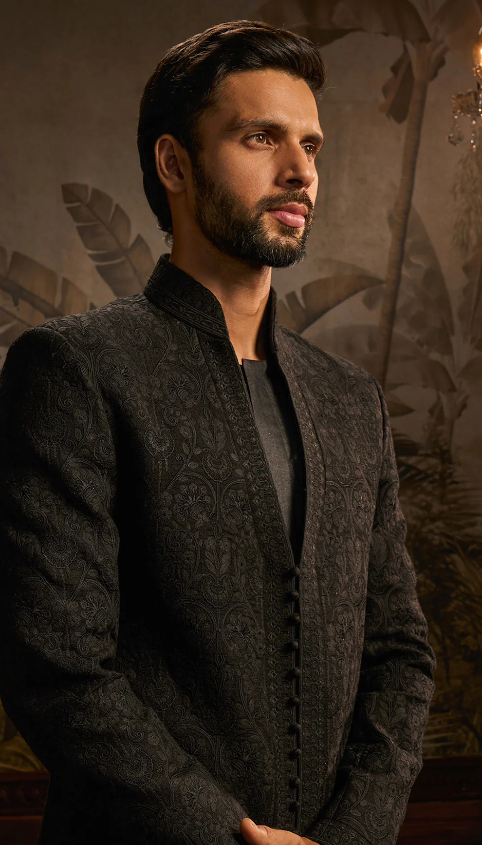 Man in a black jacket by designers Shyamal and Bhumika standing in a room with decorative wall art.